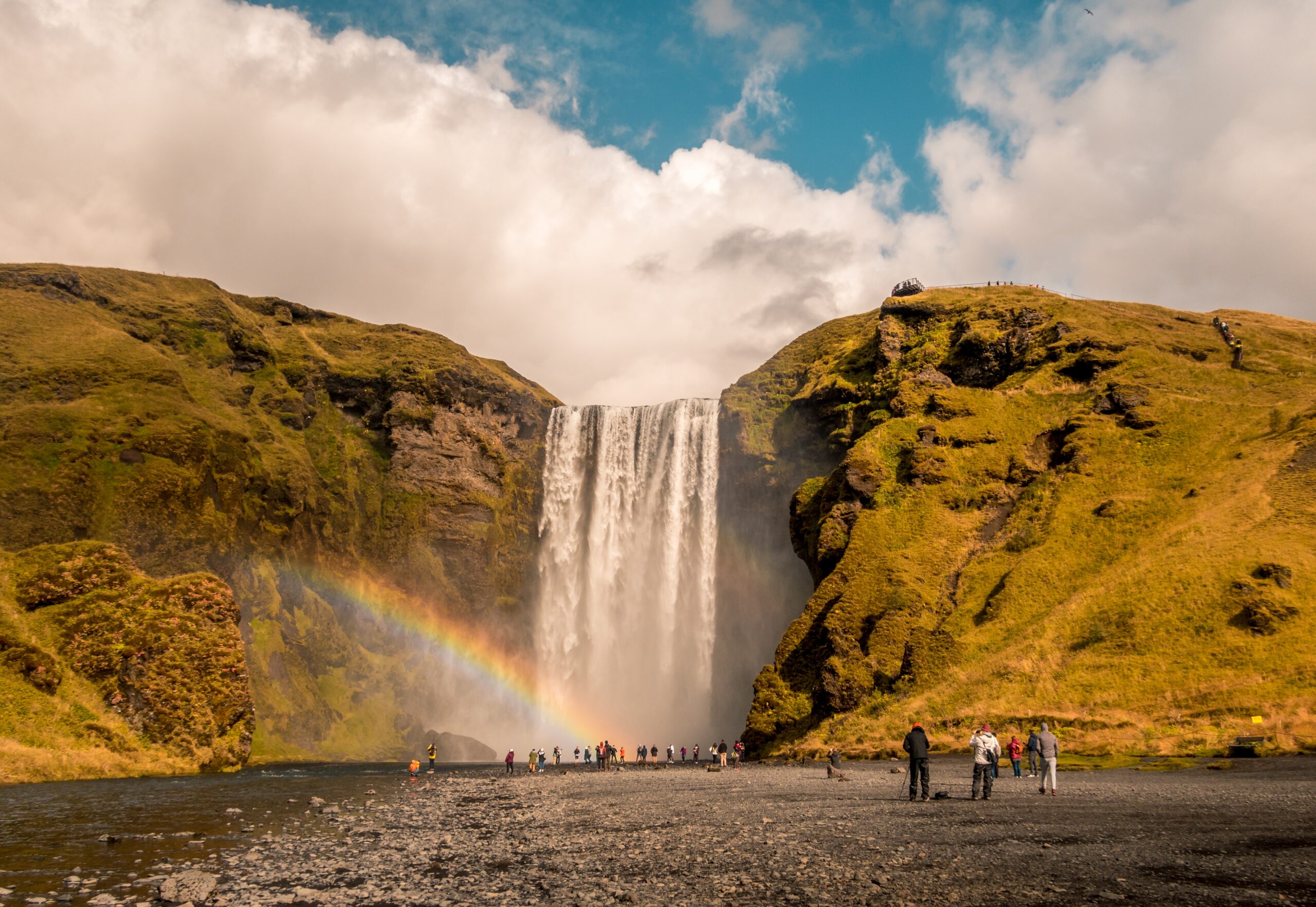 A beautiful shot of people standing near the waterfall with a rainbow on the side in Skogafoss Iceland